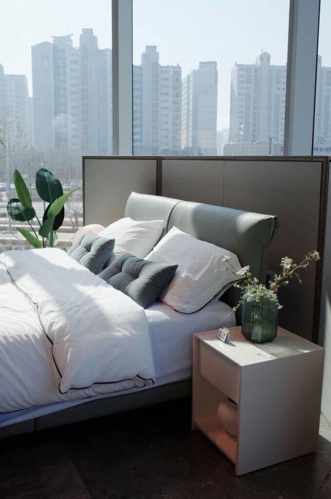 Modern bedroom with a sleek upholstered bed, white bedding, and a nightstand, featuring city views.