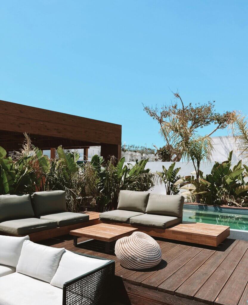 Outdoor patio with modern seating, wooden decking, lush greenery, and a pool under a clear blue sky.